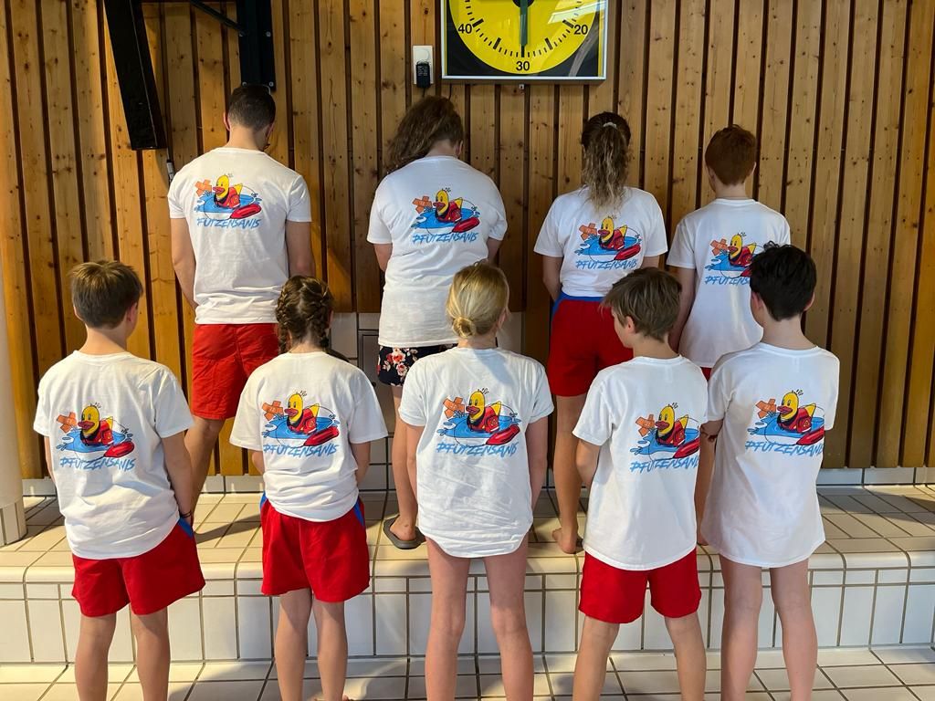 Neun Mitglieder der Wasserwacht Schliersee präsentieren im Schwimmbad die neuen T-Shirts mit dem Pfützensanis Logo.