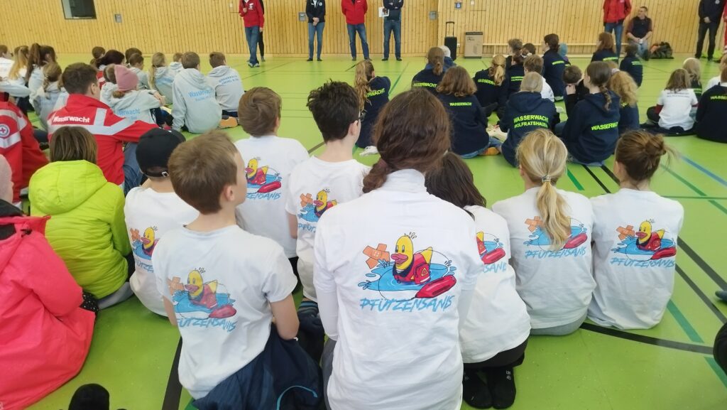 Jugendmitglieder der Wasserwacht Schliersee mit Pfützensani-T-Shirts sitzen am Boden einer Turnhalle und hören der Begrüßung zu